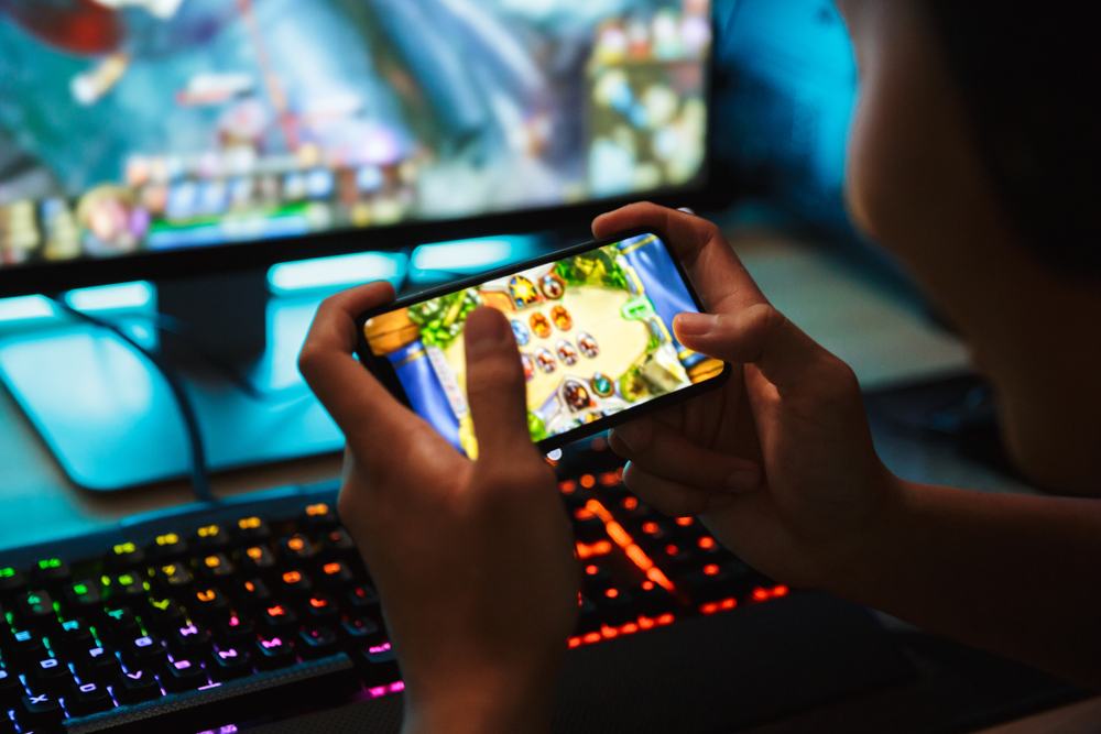 picture of a computer displaying a gameplay and a person playing another game in their cellphone